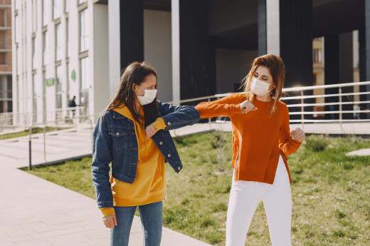 Young women in medical masks greeting each other in city 